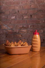 Por&ccedil;&atilde;o de coxinha sobre mesa de madeira ao lado de pote de pimenta com fundo r&uacute;stico de tijolos