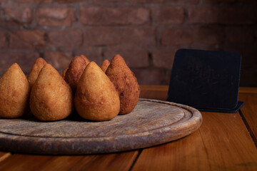 Por&ccedil;&atilde;o de coxinhas sobre mesa de madeira com placa preta para informa&ccedil;&otilde;es de valor e pre&ccedil;o com fundo r&uacute;stico de tijolos