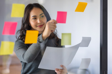 Business female employee with many conflicting priorities arranging sticky notes commenting and brainstorming on work priorities colleague in a modern office.