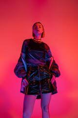 low angle view of woman in leather mini skirt looking at camera in neon light on coral and pink background.