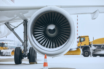 Winter day at airport during snowfall. Snowy jet engine of airplane. Snowplows clearing snow from...