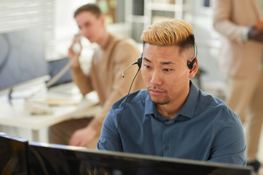 Portrait of young Asian man wearing headset in office and looking at computer screen while working in customer support or surveillance, copy space - Powered by Adobe