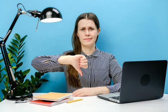 Portrait Of Disappointed Upset Unhappy Employee Girl 20s Old Years Sit Work At White Office Desk With Laptop Show Thumb Down Dislike Gesture, Posing Isolated Over Blue Color Background Wall In Studio