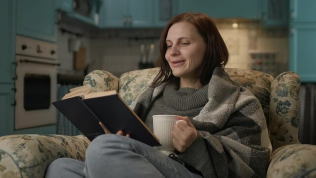 Woman reading paper book at home. 30s female enjoys reading literature, drinking hot tea sitting on cozy couch.