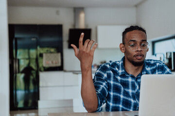 African American man in glasses sitting at a table in a modern living room, using a laptop for business video chat, conversation with friends and entertainment