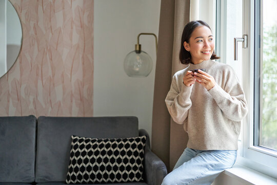 Portrait Of Young Asian Woman Sitting Near Window And Looking Outside, Drinking Hot Coffee From Espresso Cup And Enjoying Her Cozy Day Off At Home