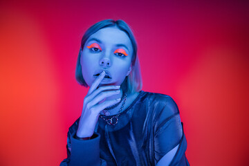 sensual young woman with neon makeup touching lips in blue light on coral and pink background.