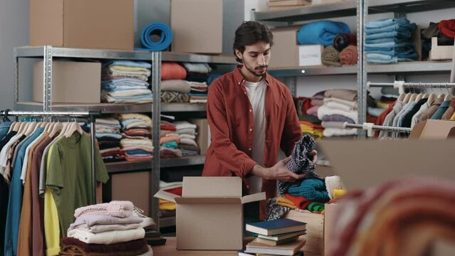 Multicultural male volunteer selecting clothes for recycling or donating and putting it into the cupboard box at the charity shop. Reuse, second-hand, conscious consumer, sustainability concept