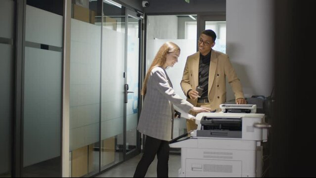 Two young multiethnic colleagues is copying documents at office standing near copier machine 