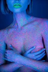 cropped view of nude woman with green neon lips and bright paint splashes on body covering breast isolated on dark blue.
