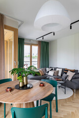 Creative composition of dining room with living room interior with wooden round table, chairs, grey sofa, pillows, gold vase with leaves, green curtain and personal accessories. Home decor. Template.