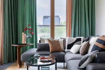 Cozy living room interior with modern gray sofa, pillows, glass coffee table, green curtain, big...