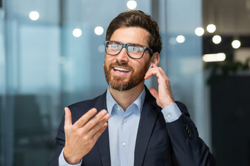 Close-up photo. A young man in the office and talking through a headset through his hands free. He is holding an earphone with his hand, gesturing with his hand, smiling.