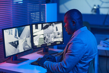 Side view portrait of black man looking at surveillance camera footage in security center office,...