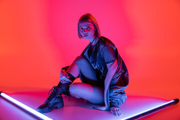 young and fashionable woman sitting near neon lamps and looking at camera on purple and coral...