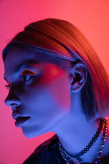 portrait of young woman with necklaces and neon makeup in blue light on pink and coral background.