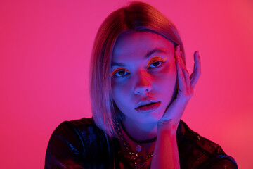 portrait of pretty woman with neon makeup holding hand near face in blue light on deep pink background.