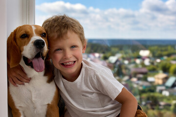 Joyful handsome boy sits against the background of window hugging his red pet. Kind dog of the Beagle breed allows himself to be hugged by a small child friend