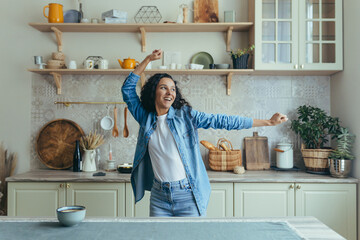 Young beautiful woman at home singing and dancing in the kitchen, cheerful Latin American woman having fun alone with curly hair.