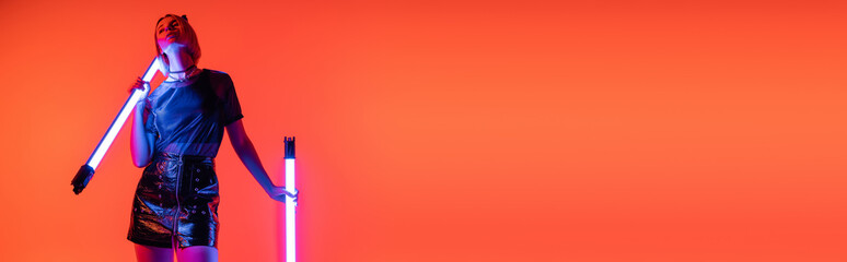 woman in stylish clothes holding glowing neon lamps while standing on coral background, banner.