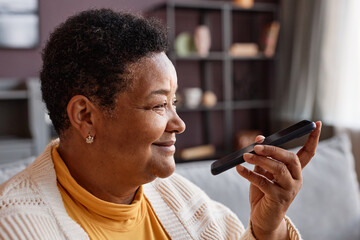Side view portrait of smiling black senior woman recording voice message via smartphone at home, copy space