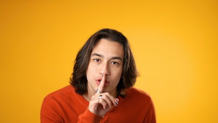 Portrait of Hispanic Latino gender fluid young hipster man 20s looks around put finger to lips shh quiet isolated on yellow background studio portrait