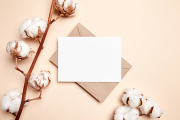 Holiday greeting card mockup with dried branch of cotton flowers and envelop on beige background, top view, flat lay. White wedding invitation card mock up
