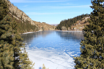 Alpine lake Kolsai covered with ice with a distant view of the mountain gorge, there is snow and traces on the ice in some places, spruce trees in the foreground, winter, sunny weather