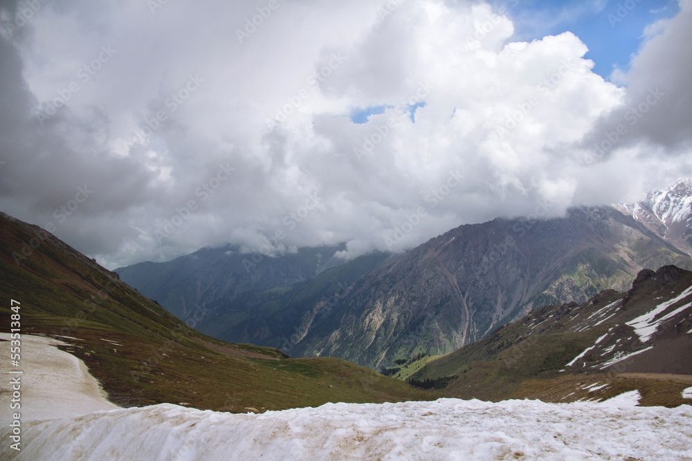 Sticker Mountain range with peaks, thick clouds on the peaks, sunlight on the slopes, snow in the foreground, Chimbulak mountain resort, winter