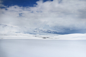 White snow-covered Assy plateau with mountain snow-covered ridges in the background, sunlight in the middle of the plateau and on the ridges, beautiful thick clouds, winter