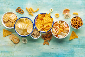 Salty snacks. Party food mix. Potato and tortilla chips, crackers and other appetizers in bowls, overhead flat lay shot on a blue background