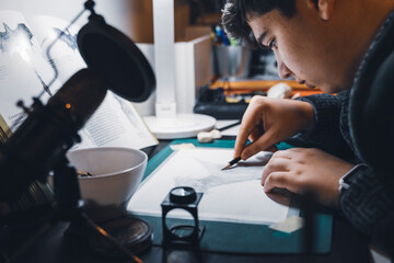 Close-up of a young man drawing at his study table. Young man holding a pencil and drawing something, warm orange light at home and a microphone for podcasts and social networking.