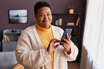 Waist up portrait of black senior woman holding smartphone standing by window at home and looking...