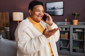 Waist up portrait of cheerful senior grandma speaking on phone in home setting and gesturing