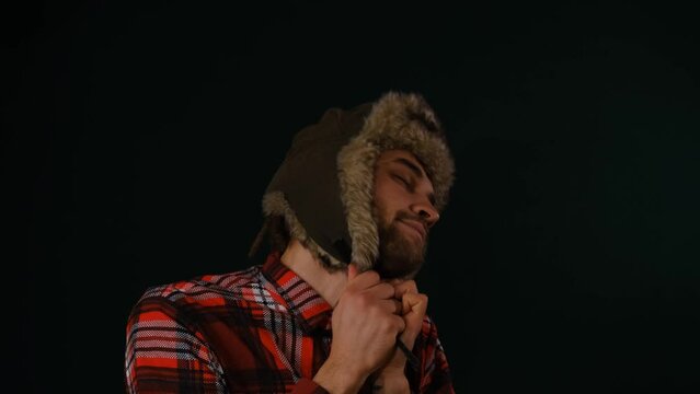 Young Caucasian bearded man wears fluffy fur hat with earflaps on head and dances on dark studio background. Classic Russian stereotype simple Siberian guy in national clothes of Northern peoples.