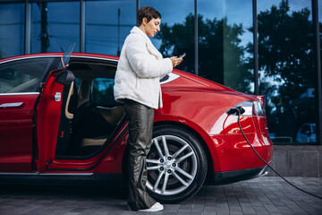 Woman using phone while charging electric car by the office building