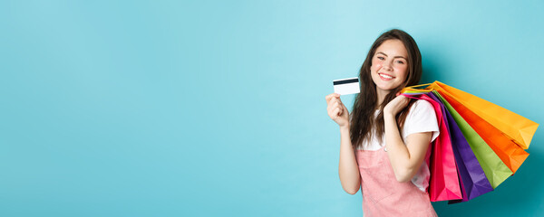 Young satisfied woman smiling, showing plastic credit card and holding shopping bags, buying with...