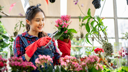 Happy young adult asian beautiful gardener florist woman in gloves and apron. Plants flowers in greenhouse  home garden. Gardening and floriculture. Young enterprenuers farm modern owner.