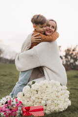 cute baby (son) gives a gift box and bouquet of flower to his beautiful blonde mother in the park on a background of green grass . mother's day concept