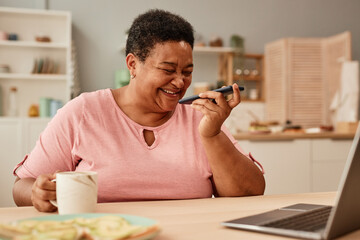 Portrait of black senior woman laughing happily while listening to voice message via smartphone
