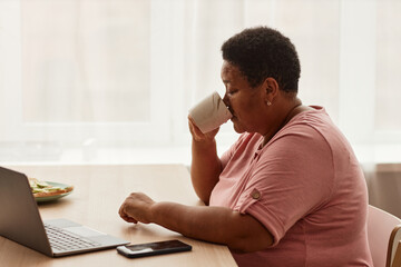 Side view portrait of black senior woman enjoying cup of coffee at breakfast and using laptop