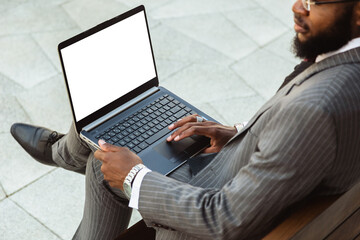 A dark-skinned man in a business suit is using a laptop outdoors. Working break. Online learning and education. Distance courses and communication. Copy space and mock up