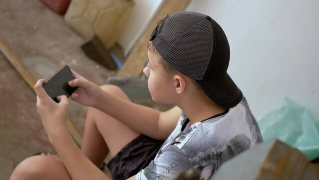 Teenager Playing Video Games on Mobile Phone while Sitting on Chair in Garage. Top view. A boy in a cap looks at the screen of a mobile phone, playing, relaxing in a secluded place in sun's rays.