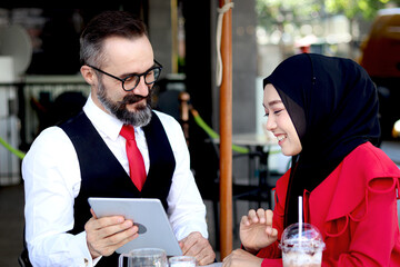 Senior elderly mature business man using tablet and discussing with hijab Muslim women together during having a coffee at outdoor coffee shop, happy harmony people take a break from drinking beverage