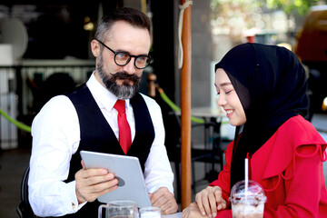 Senior elderly mature business man using tablet and discussing with hijab Muslim women together during having a coffee at outdoor coffee shop, happy harmony people take break from drinking beverage