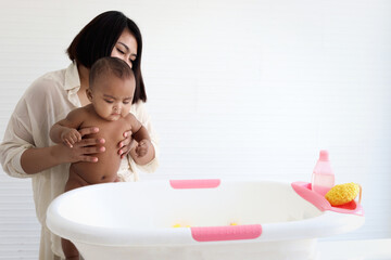 Obraz na płótnie Canvas Mother holding six month Asian African American infant baby to take bubble bath in bathroom, mother bathing kid in tub, mom washing and cleaning her child in bath.