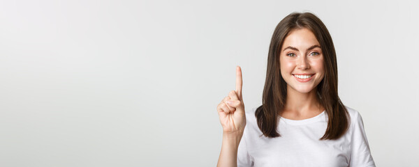 Naklejka na ściany i meble Close-up of cheerful attractive young woman smiling, showing one finger, white background