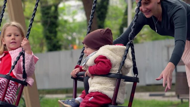 fun, playground - young smiling mom pushes her children on the swing at park