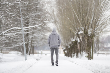 Young adult man slowly walking on white snow covered sidewalk at park in cold winter day. Spending time alone. Enjoying peaceful stroll. Back view.