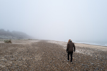 Lone woman seen walking along a Suffolk beach during mid winter. Dense fog has started to roll in from the North Sea.